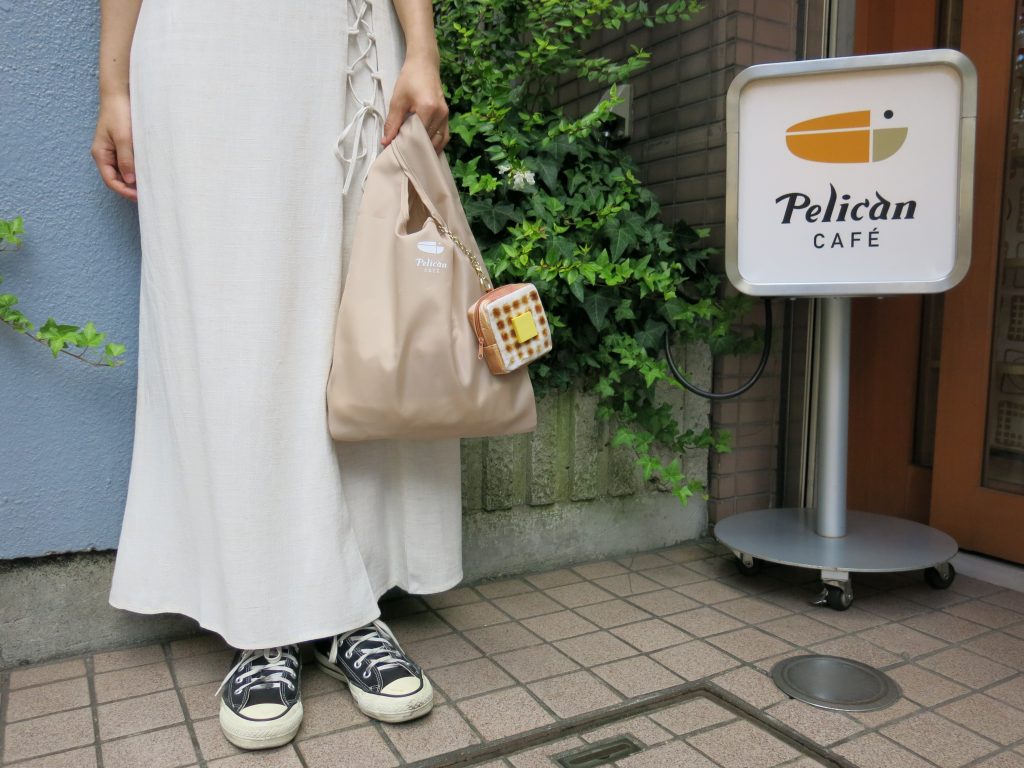 ペリカンカフェの看板と、エコバッグにポーチをつけて立つ女性